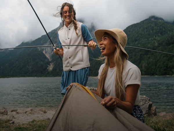 Two women in a mountain landscape, laughing while erecting a tent.